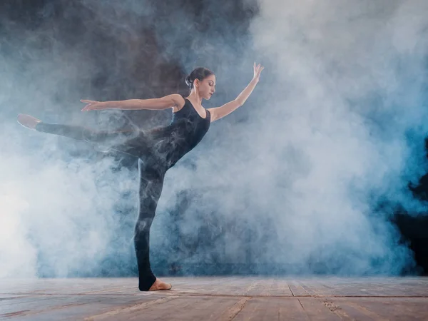 Jovem bela bailarina no palco de fumaça dançando balé moderno. executa movimentos suaves com as mãos contra holofotes de fundo. Mulher de traje preto em cena — Fotografia de Stock