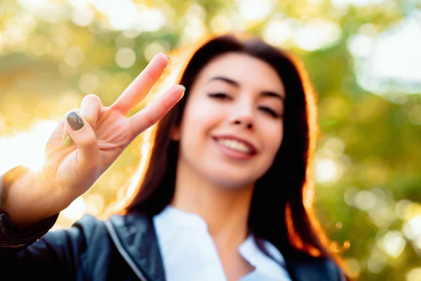 Молодая красивая женщина показывает жест МИРА и улыбается в камеру на фоне осеннего парка . — стоковое фото