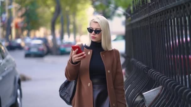 Retrato de una joven rubia atractiva usando un dispositivo móvil en la ciudad de otoño. Chica tienen un aspecto elegante, gafas de sol y piercing en la nariz. Dama caminando sola por la calle. Tecnología — Vídeo de stock