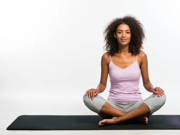 Mujer afroamericana feliz en ropa deportiva cómoda sentada en alfombra de yoga negro sobre fondo blanco de la pared. Chica concentrada en su práctica — Foto de Stock