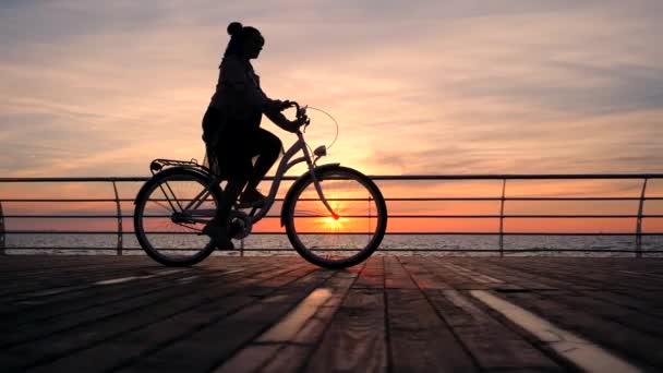 Nádherný východ nebo západ slunce nad oceánem. Silueta dívka stylové cyklistické na vintage kolo na dřevěných nábřeží. Žena na kole poblíž moře. Zpomalený pohyb. 4k.