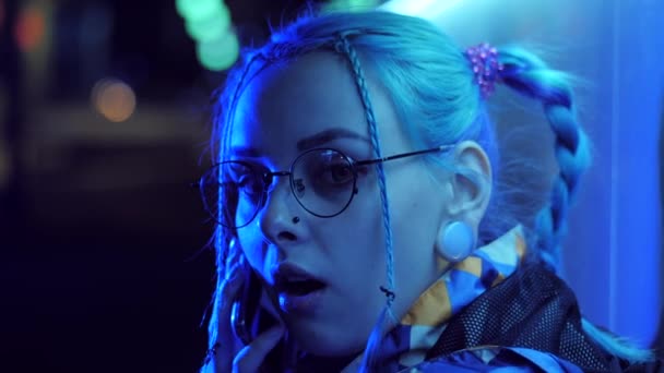 Attractive pretty girl with unusual hairstyle near glowing neon lights of the city at night talking with smartphone. Dyed blue hair in braids. Happy hipster teenager using mobile — Stock Video