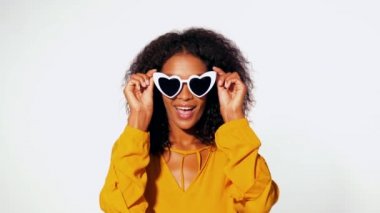 Sarı top kalp giyen afro saçlı mutlu Afrikalı-Amerikalı kadın trendy güneş gözlüğü beyaz duvar arka plan üzerinde şeklinde. Flört, kameraya gülümseyen kız eğleniyor.