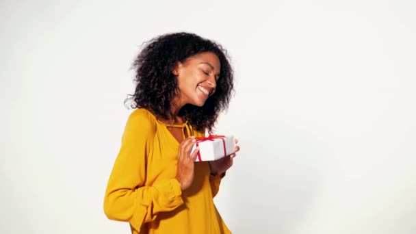 Carino afro-americano giovane donna in giallo autunno top contenente confezione regalo con nastro rosso e fiocco su sfondo bianco parete. Ragazza sorridente, lei è felice di ottenere presente . — Video Stock