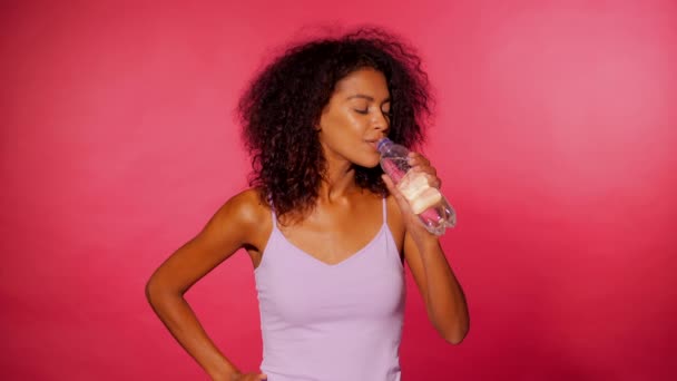 매력적인 흑인 스포티 한 젊은 여자 핑크 벽 바탕에 물 한 병을 마시는. 피트 니스 후 스포츠 옷에서 소녀입니다. 스튜디오 영상입니다. 4 k — 비디오