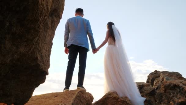 Unrecognizable wedding couple holding hands on sunset or sunrise background. Bride and groom standing outdoor near sea. Sun flares. Slow motion — Stock Video