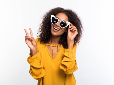 Sarı aşınma ve kameraya gülümseyen ve Tamam jesti beyaz duvar arka plan üzerinde gösterilen güneş gözlüğü afro saçlı güzel Afrikalı-Amerikalı kadın. Karışık yarış kız portre.