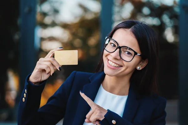 Successful pretty woman look to camera and showing unlimited gold credit card on office building background. Professional female manager wearing glasses and suit