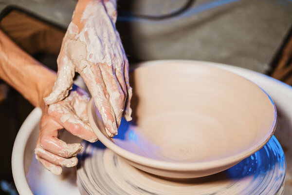 Experienced potter shapes the clay product - plate - with pottery tools. Close up of male hands working on potters wheel. Shot of half-finished ceramic plate spinning on jigger