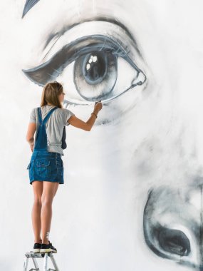 Güzel kız kentsel sokak duvarında büyük kadın yüzü aerosol sprey ile graffiti yapmak. Yaratıcı sanat. Resim çizim denim tulum yetenekli öğrenci