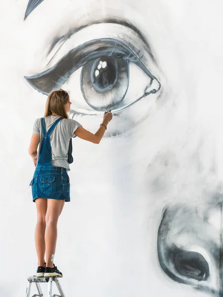 Beautiful girl making graffiti of big female face with aerosol spray on urban street wall. Creative art. Talented student in denim overalls drawing picture — Stock Photo, Image