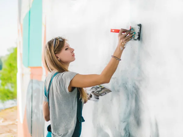 Beautiful Young Blonde Girl making graffiti of big eye with aerosol spray on urban street wall. Creative art. Talented student in denim overalls drawing picture — Stock Photo, Image