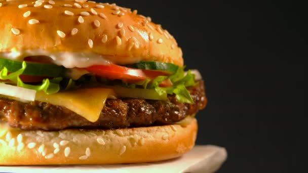 Hambúrguer apetitoso grande com costeleta de carne, cebola, legumes, queijo derretido, alface e molho de maionese. Hambúrguer isolado gira sobre fundo escuro, vista close-up . — Vídeo de Stock