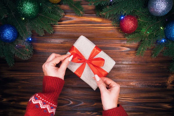 Las manos femeninas en el jersey caliente rojo corrige el lazo rojo por presente en el papel plateado. Mesa vintage de madera con decoraciones navideñas. Vista del plan superior. El humor de fiesta, el concepto de año nuevo . — Foto de Stock