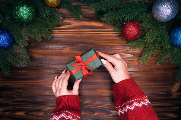 Las manos femeninas ponen regalo envuelto en papel esmeralda con cinta roja en la mesa de madera retro con decoraciones de Navidad ramas de abeto verde, bolas de Navidad. Vista superior del plan . — Foto de Stock