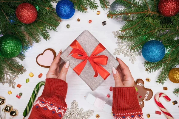 Mani in maglione caldo contenente regalo in carta argentata con fiocco rosso. Tavolo bianco in legno con decorazioni natalizie - rami di abete rosso con palline di Natale, pan di zenzero, lecca-lecca . — Foto Stock