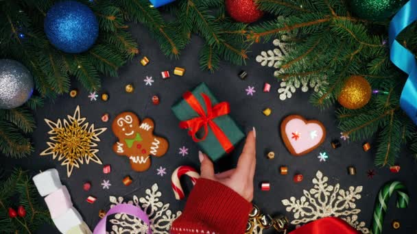 Female hands puts gift wrapped in emerald paper with red ribbon on monochrome surface and then takes it. Black table with Christmas decorations - gingerbread, snowflakes, lollipops.Top plan view — Stock Video