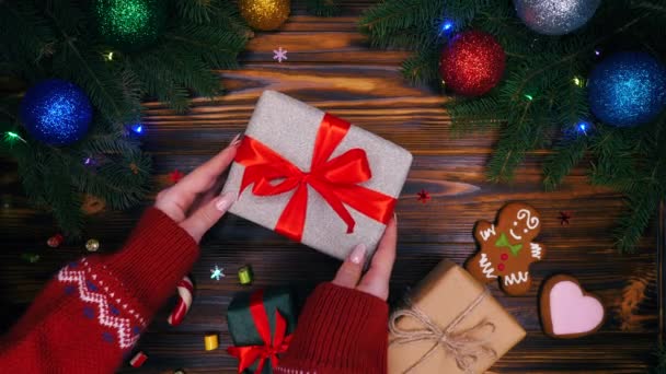 Le mani mettono il regalo avvolto in carta argentata sulla superficie di legno e poi lo prendono. Tavolo con decorazioni di Natale - pan di zenzero, scatole presenti. Vista dall'alto. Concetto vacanze . — Video Stock
