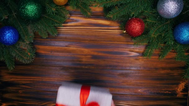 女の子の手はモミの枝で作られたフレームの中央にシルバーの紙で包まれた贈り物を置くし、それを引き継ぎます。クリスマスの装飾 - つまらないものガーランド ライトが点滅します。トップ ビュー — ストック動画