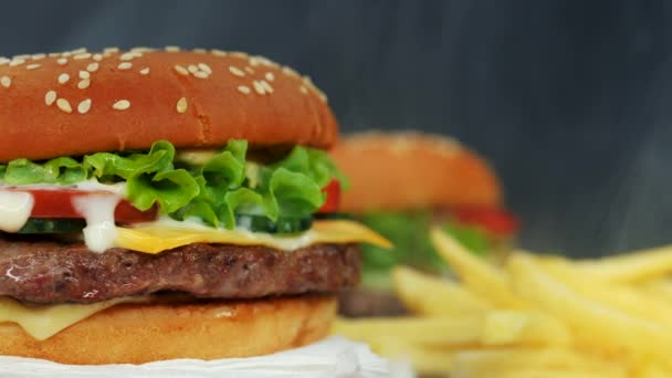 Hamburguesas a la parrilla caseras frescas y papas fritas girando sobre fondo de humo negro. Patty carne, tomates, pepino, lechuga y semillas de sésamo. Delicioso concepto de comida rápida — Vídeos de Stock