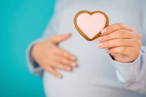 Έγκυος γυναίκα κρατώντας μπισκότο σε σχήμα καρδιάς σε κοιλιά κοιλιά φόντο. Νεαρό κορίτσι σε μπλε φορώντας περιμένοντας μωρό. Μητρότητας, μητρότητας, εγκυμοσύνη, έννοια αγάπης. — Φωτογραφία Αρχείου