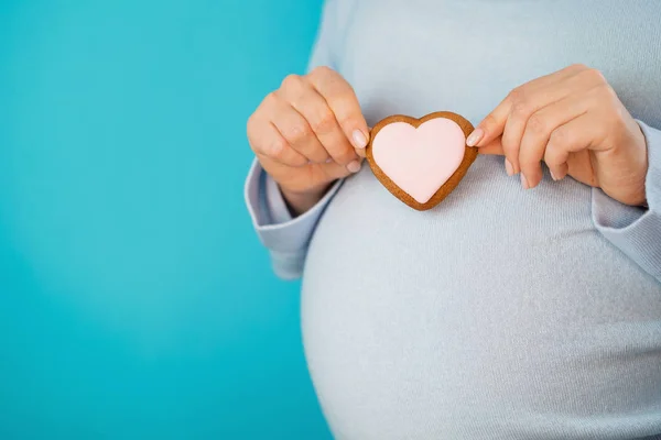 Έγκυος γυναίκα κρατώντας μπισκότο σε σχήμα καρδιάς σε κοιλιά κοιλιά φόντο. Νεαρό κορίτσι σε μπλε φορώντας περιμένοντας μωρό. Μητρότητας, μητρότητας, εγκυμοσύνη, αγάπη έννοια. Χώρο αντίγραφο — Φωτογραφία Αρχείου