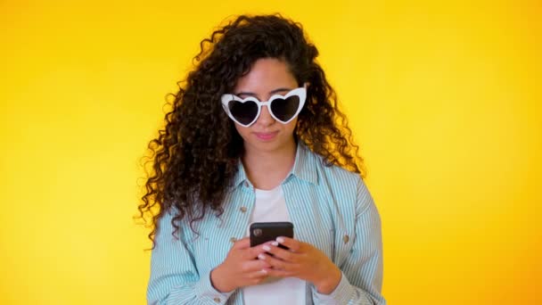若い女性の笑顔と黄色の背景上の携帯電話を使用しています。美しい混合レースの女の子を保持し、スマートフォンでテキストメッセージ。技術を持つ女性. — ストック動画