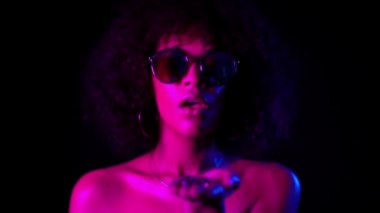 Neon ışıkta siyah arka planda konfeti üfleme kıvırcık saçlı Genç Afrikalı kız. Kutlama yapan kadın, sevinç ve mutluluğu tasvir ediyor. Başarı, zafer, tatil konsepti.