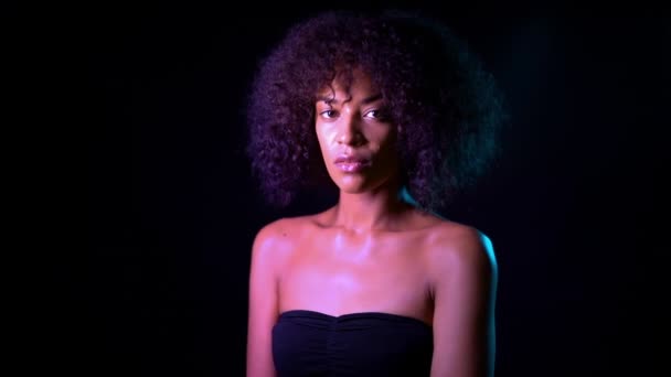 Retrato de una joven afroamericana seductora en luz de neón rosa sobre fondo negro. Mujer tentadora con maquillaje perfecto mirando a la cámara y sonriendo. Glamour, concepto de moda . — Vídeos de Stock