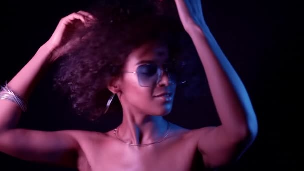 Chica africana joven con el pelo rizado bailando en luz de neón. Mujer tentadora con maquillaje perfecto sonriendo, disfrutando del momento, de la música. Glamour, moda, concepto de estilo — Vídeos de Stock