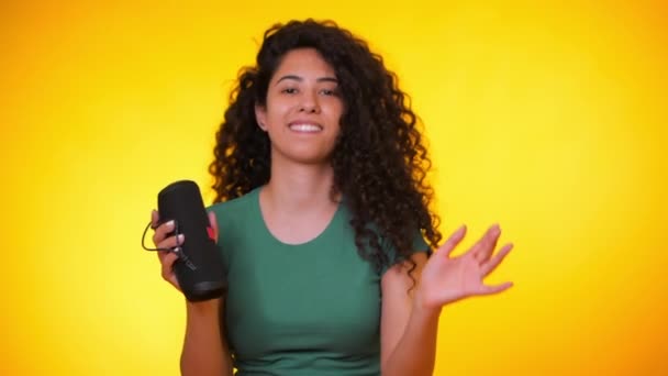Modern trendy girl listening to music by wireless portable speaker. Young beautiful woman with curly hairstyle enjoying and dancing at yellow background. Female moves to the rhythm of music. — Stock Video