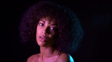 Neon ışıkta karanlık arka planda kıvırcık Afrika saçlı karışık yarış kız. Makyaj ve giyim olmadan kamera bakarak seksi kadın. 4k.