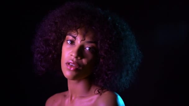 Mixed race girl with curly african hair on dark background in neon light. Sexy woman with make-up and without clothing looking at camera. 4k. — Stock Video