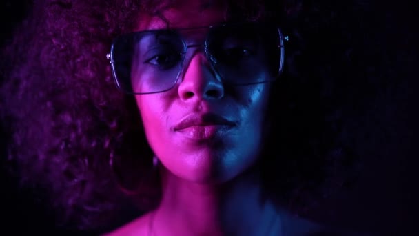 Retrato de una joven mulata en luz de neón violeta. Moda, glamour, concepto de modelo. Mujer seductora con maquillaje y gafas transparentes posando en habitación oscura por la noche . — Vídeos de Stock