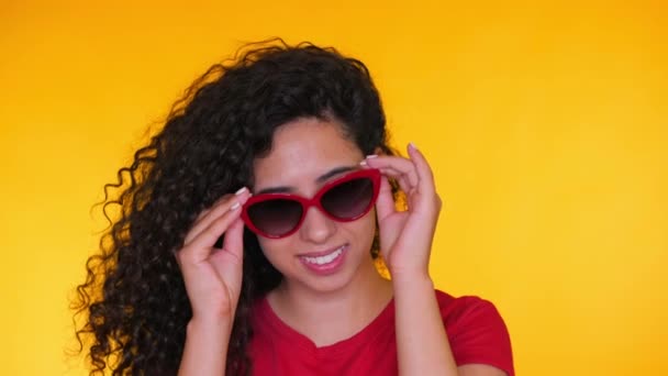 Portrait of young spanish girl with curls puts on sunglasses on yellow background. Tempting woman smiling to camera. Studio footage. Female in red t-shirt, casual style — Stock Video