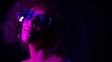 Closeup portrait of trendy black woman with afro hair in neon purple light smiling and looking at camera in studio against dark background. slow motion.