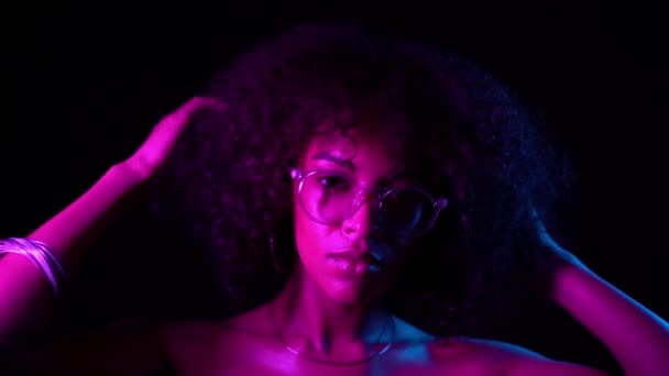 메이크업과 투명 한 안경을 가진 섹시 한 여자는 검은 배경에 유혹. 네온 빛에 젊은 매력적인 아프리카 여자의 초상화입니다. 파티, 나이트 클럽 컨셉 — 비디오