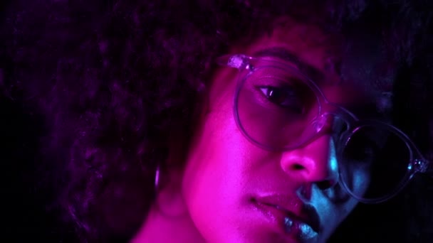 Mulher tentadora com maquiagem perfeita e óculos transparentes posando no quarto escuro à noite. Retrato de jovem menina africana sexy em luz de néon — Vídeo de Stock