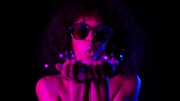 Young mulatto girl with curly hair blowing confetti on dark background in neon light. Woman celebrating, depicts joy and happiness. Success, victory, holiday, new year concept. — Stock Footage Young mulatto girl with curly hair blowing confetti on dark background in neon light. Woman celebrating, depicts joy and happiness. Success, victory, holiday, new year concept. — Stock Video