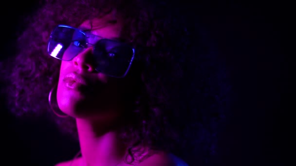 Närbild porträtt av trendig svart kvinna med afro hår i neonlila ljus leende och tittar på kamera i studion mot mörk bakgrund. långsam rörelse. — Stockvideo