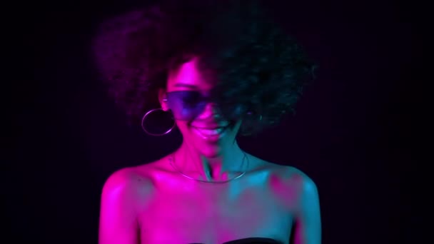 黒の背景に対してスタジオで楽しい笑顔とダンスを持つネオンパープルライトでアフロの髪とセクシーなムラートの女性の肖像画。スローモーション. — ストック動画