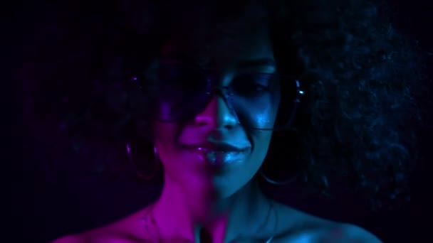 Close-up portret van mooie zwarte vrouw met Afro haar in neon paars licht met plezier glimlachen en dansen in de studio tegen zwarte achtergrond. Slow Motion — Stockvideo