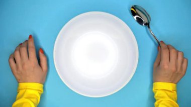 Kaşıklı genç kadının elleri. Bayan restoranda oturup sipariş bekliyor. Onu yemek için beklerken kız masada sinirle parmaklarını vuruyor. Mavi arka planda boş beyaz plaka