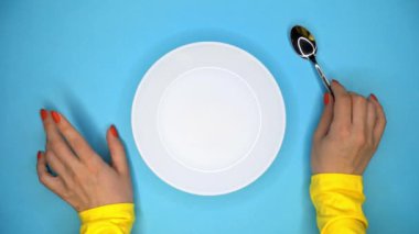 Kaşıklı genç kadının elleri. Bayan restoranda oturup sipariş bekliyor. Onu yemek için beklerken kız masada sinirle parmaklarını vuruyor. Mavi arka planda boş beyaz plaka