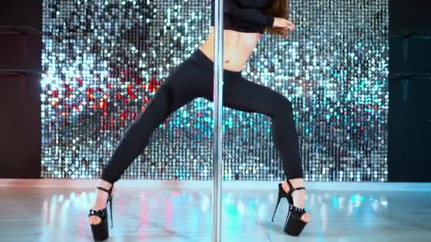 Poledance on shining wall background. Close-up legs in heels shoes of unrecognizable woman doing tricks with pylone. Young girl in black dances sexually with pole in studio — Stock Video