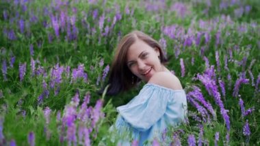 Menekşe tarlasında başını sallayan güzel saçlı bir kadın. Mavi elbiseli seksi kız gülümsüyor, flört ediyor..