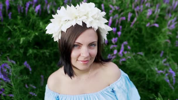 Chica en vestido de lino azul sonriendo y coqueteando. Atractiva mujer con corona de flores blancas en el campo de lavanda . — Vídeos de Stock