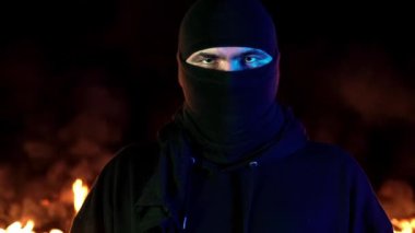 Balaclava'da genç bir adamın, yanan bir gece yangınının arka planına karşı portresi. 