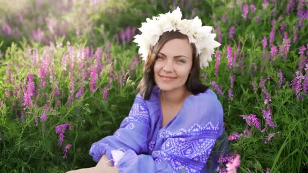 Joven mujer bonita con corona de flores sentada en el campo púrpura. Retrato de niña en ropa azul . — Vídeos de Stock