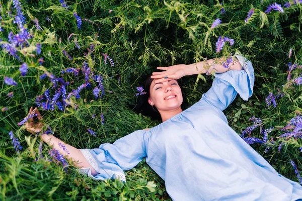 Happy smiling young woman falling down on grass with violet flowers. Beautiful girl lying in field. Enjoying nature.